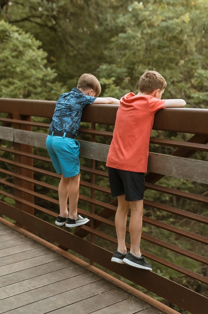 Two children in colorful shirts lean over a wooden bridge railing to look down at the forest scenery below.