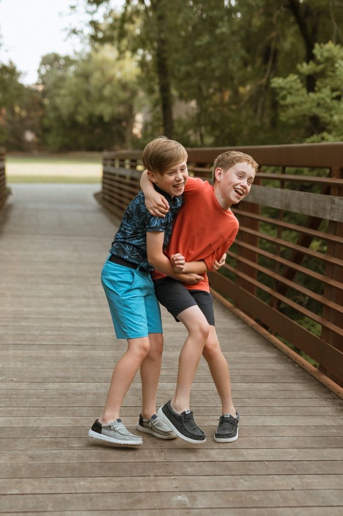 Two kids playfully hugging and laughing together on a wooden bridge walkway in a park setting.