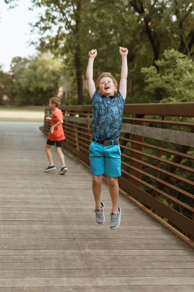 Two kids playing and jumping on a wooden bridge walkway surrounded by green trees on a summer day.