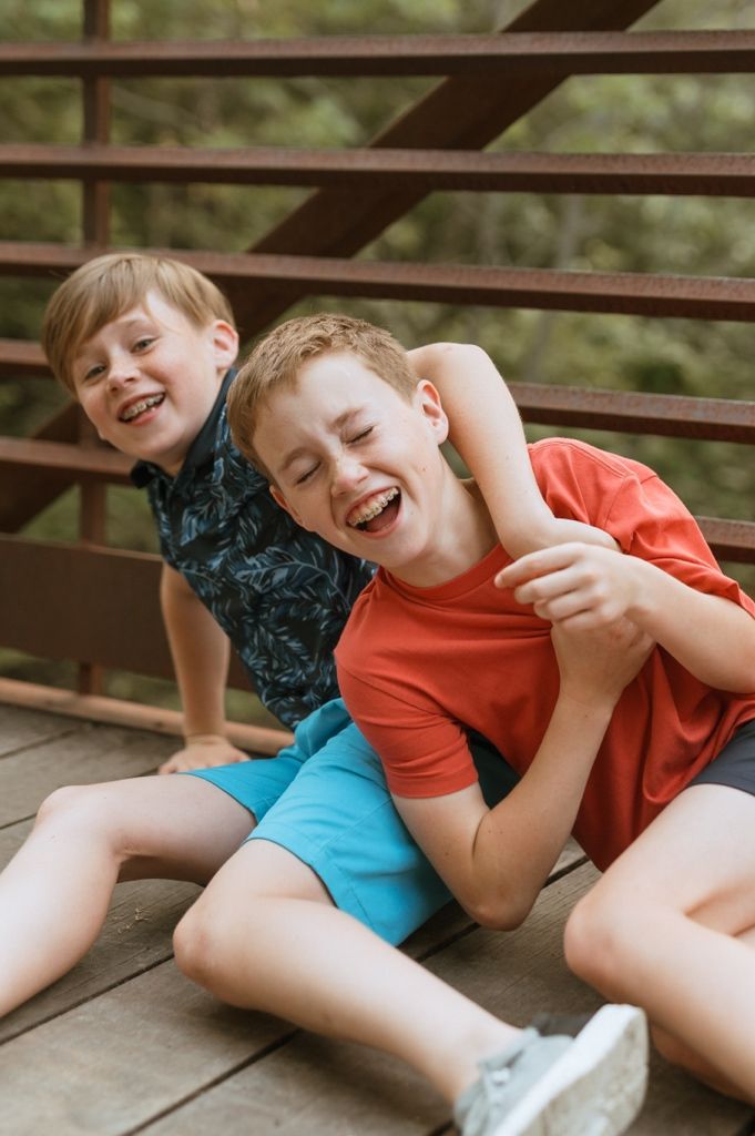 Two children laughing together while lying on wooden decking on a sunny day outdoors.