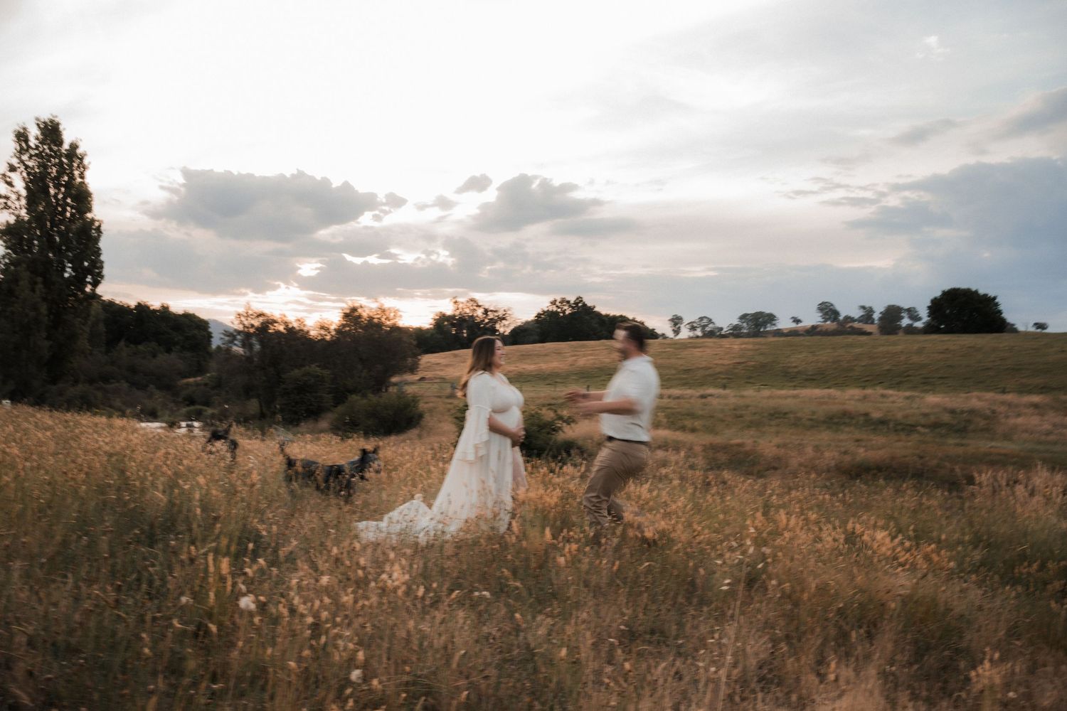 Two figures in flowing white dresses walk through a golden field at sunset with dramatic cloudy sky overhead in Mansfield, Victoria