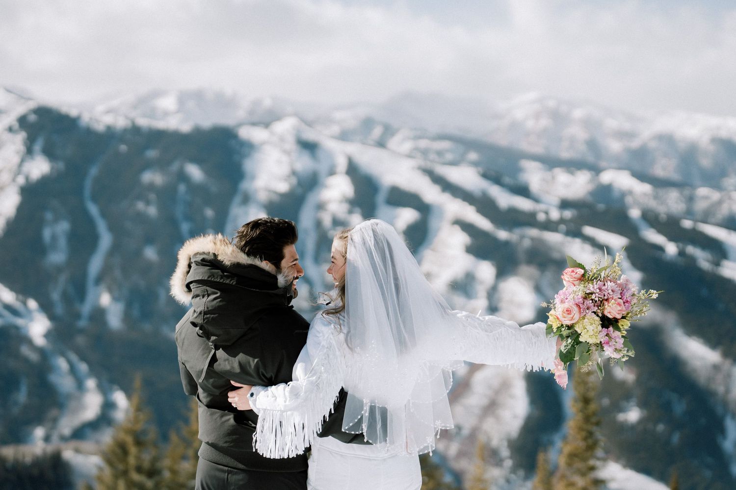 A couple embraces against a snowy mountain backdrop with pink bouquet and white veil flowing in the winter breeze.
