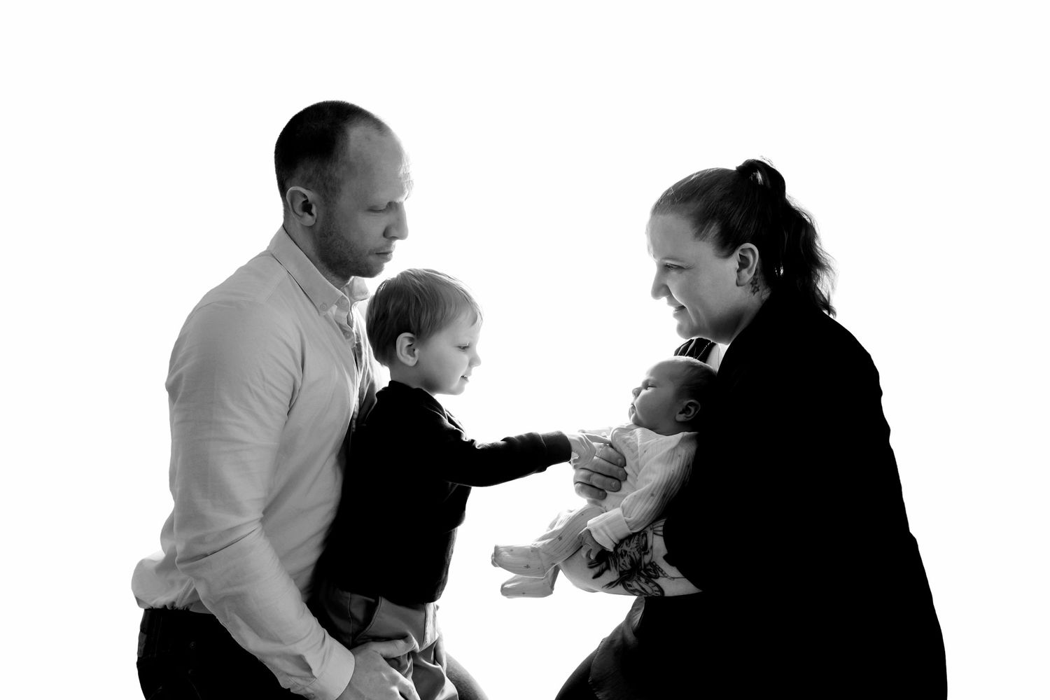 Black and white family portrait with a toddler meeting their new baby sibling while parents look on.