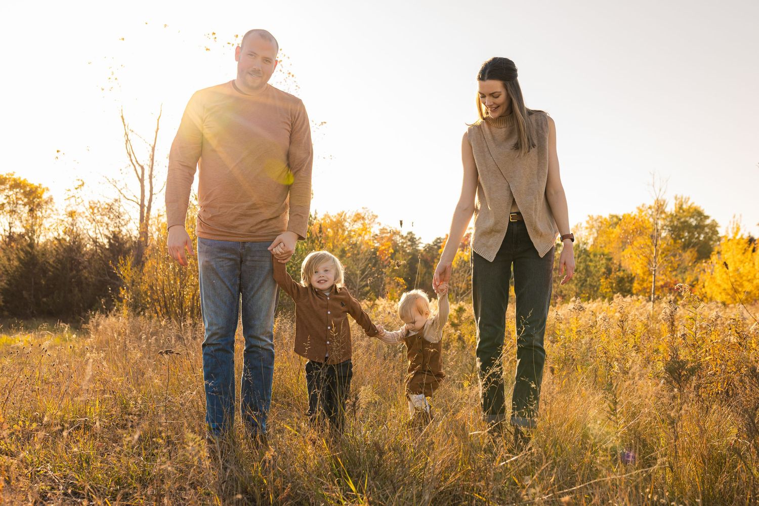 Family members walking hand-in-hand together towards the camera in a golden sunlit field during sunset.
