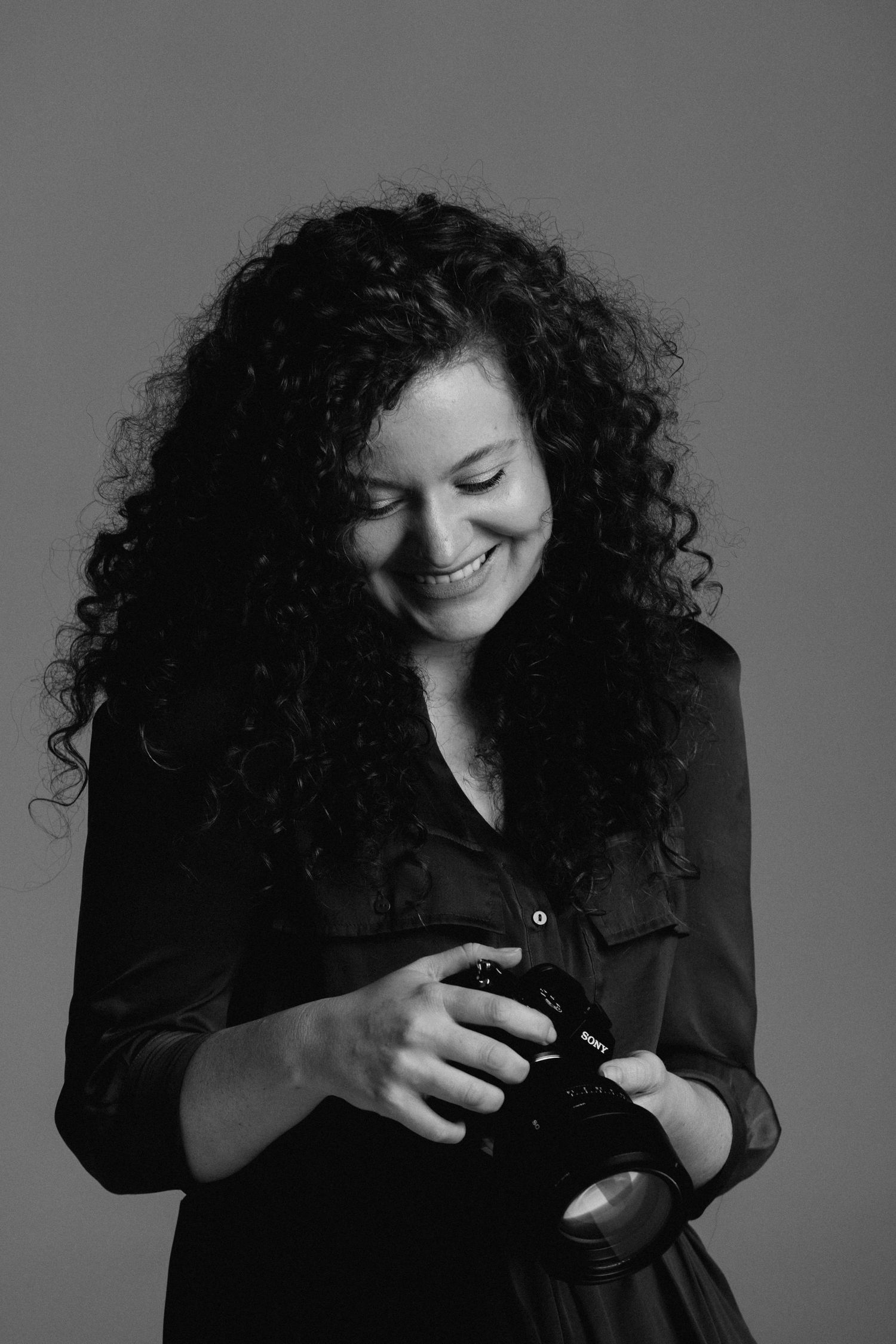 Black and white portrait of a photographer with curly hair smiling while looking down at their camera.