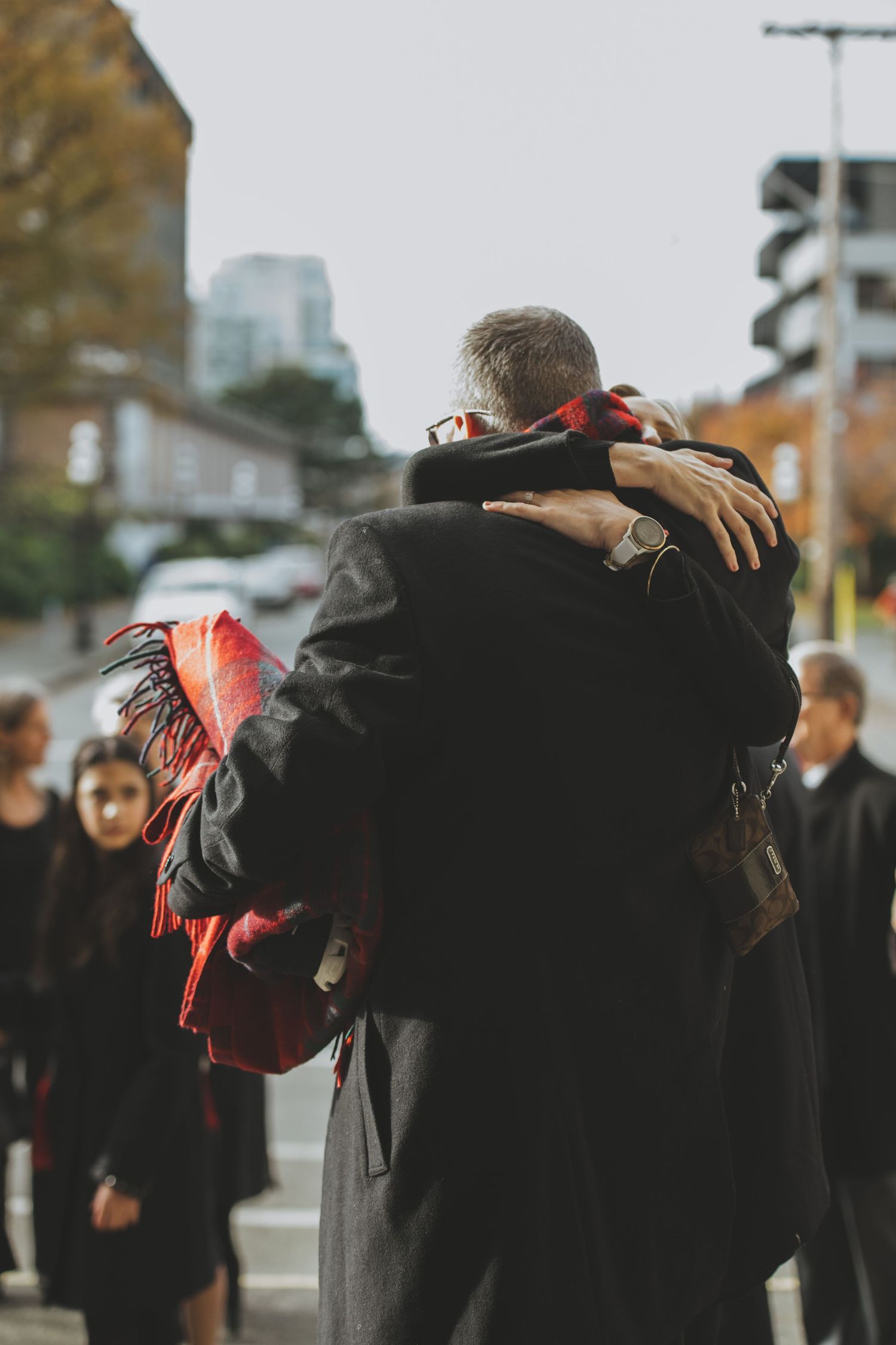 Two people embrace in a warm hug on a city street while others look on in the background.