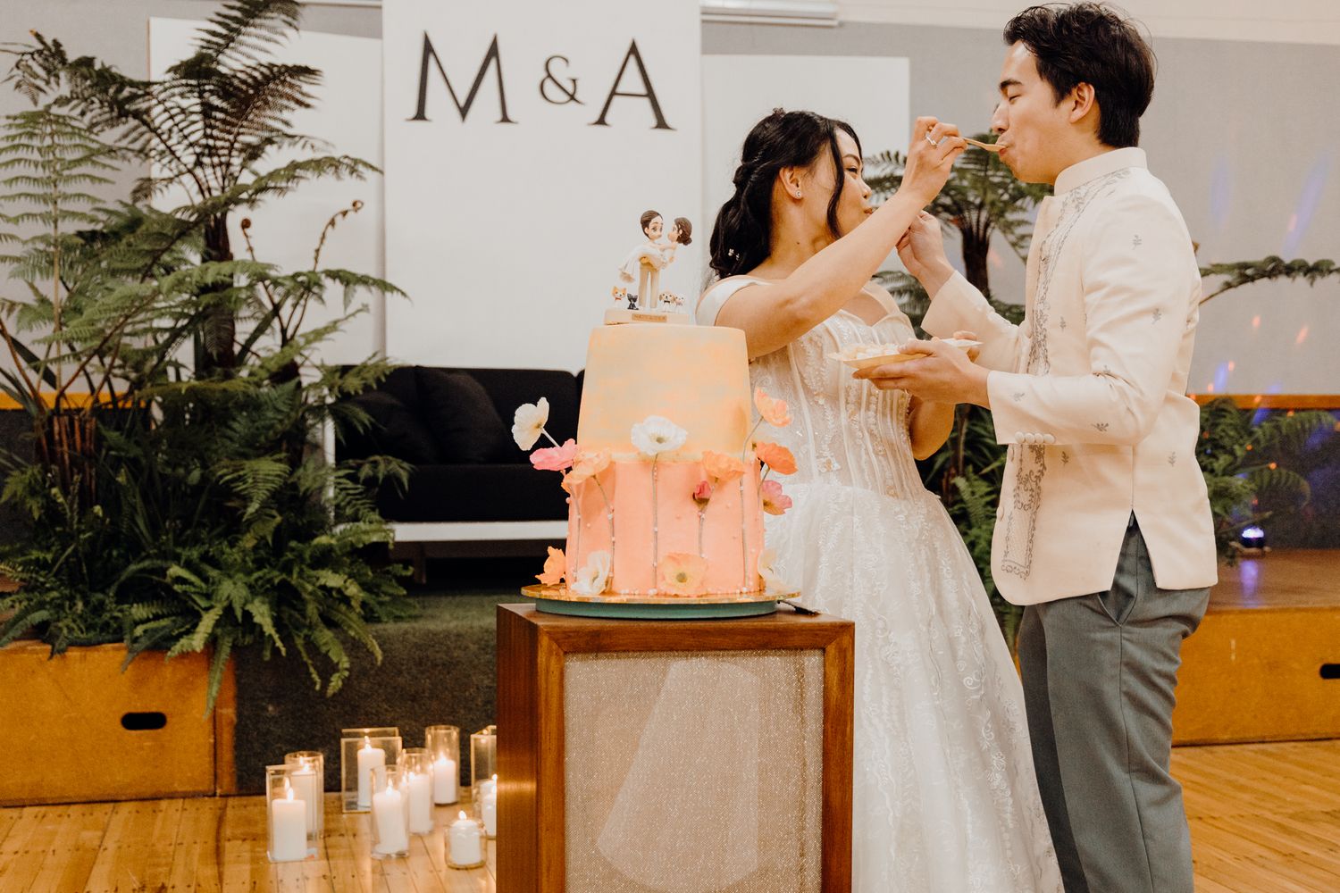 A couple shares cake during their wedding reception in front of a backdrop with M&A signage.
