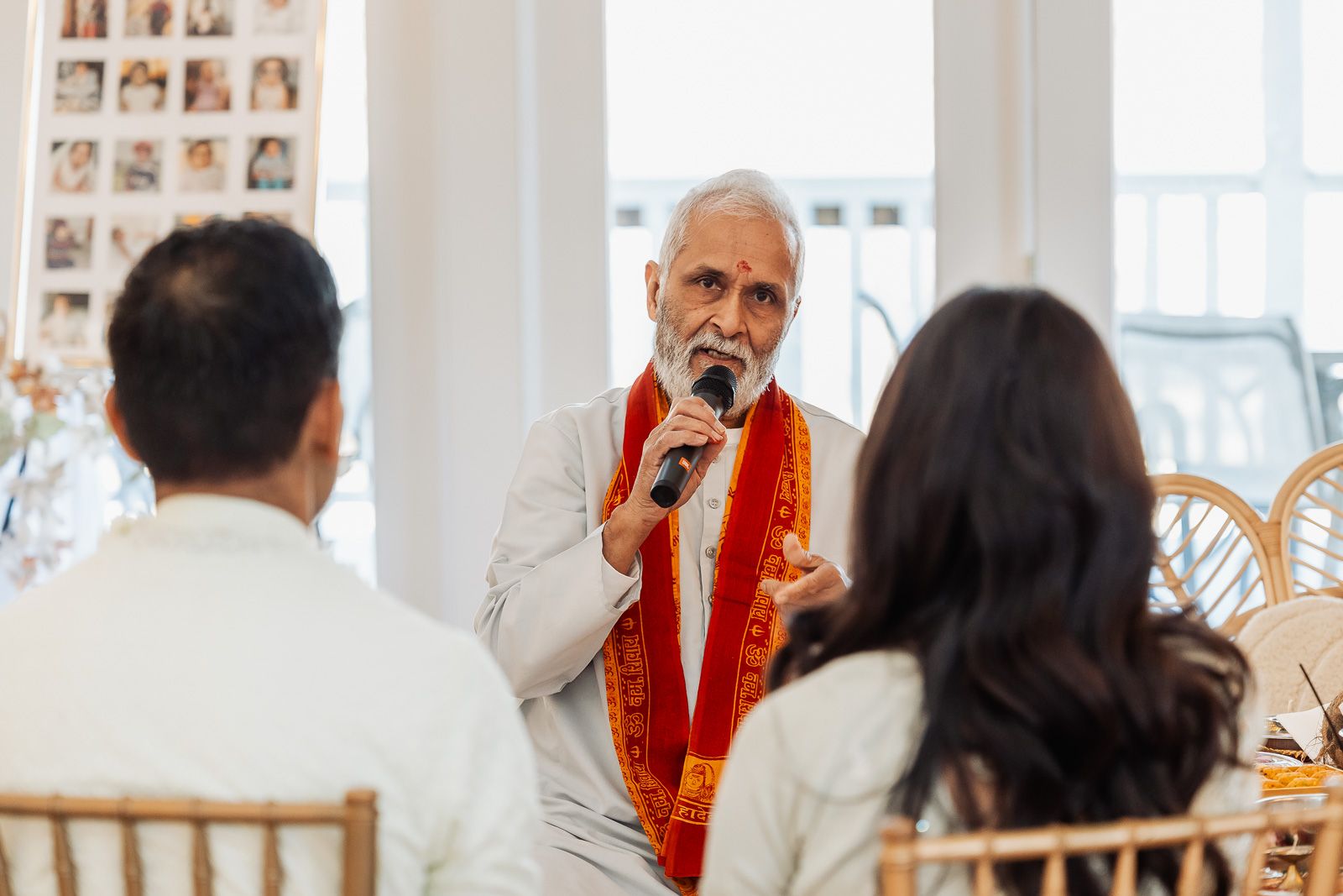 An officiant in red stole speaks into a microphone during a wedding ceremony in a bright indoor setting.