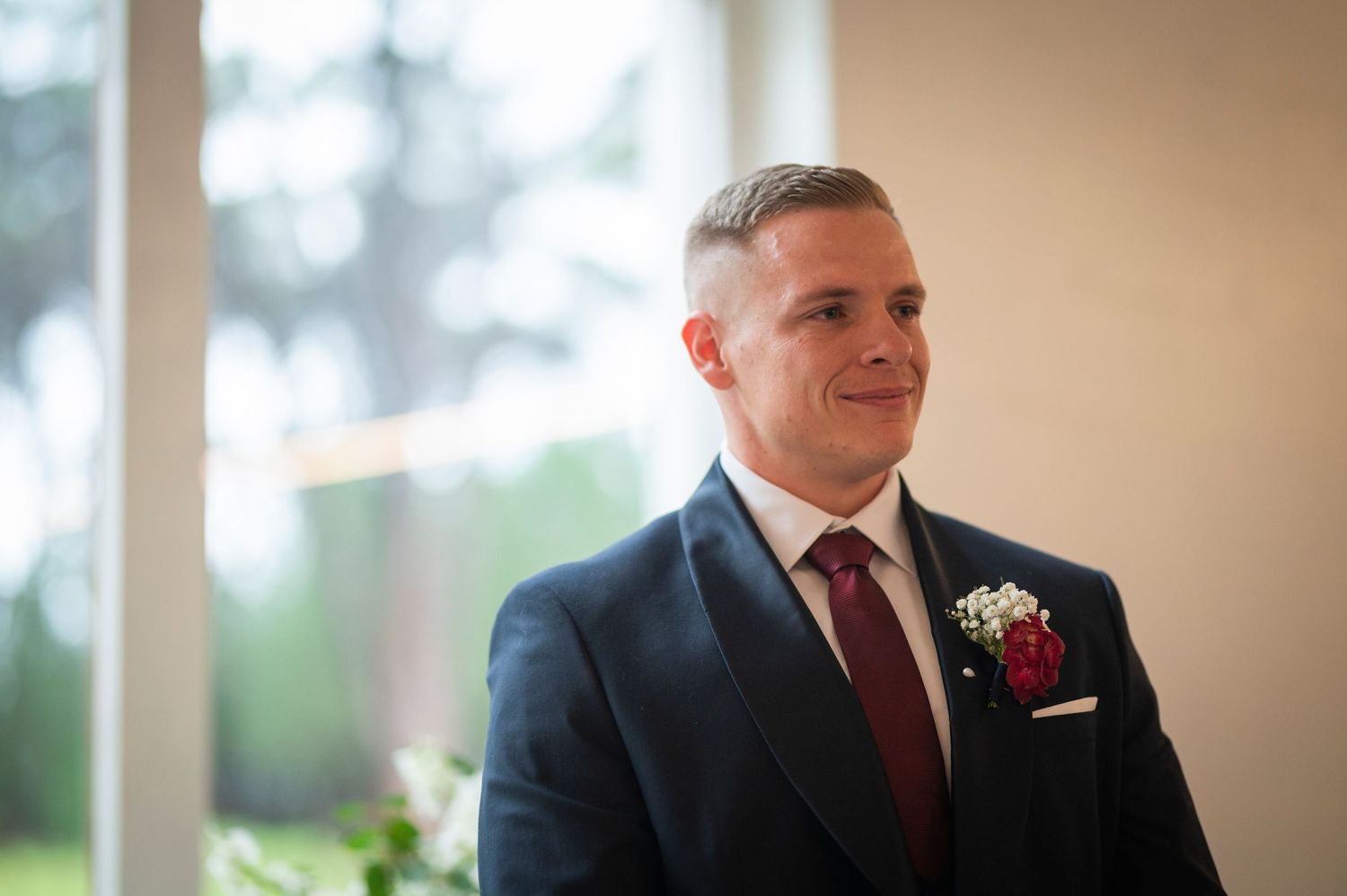 A groom in a dark navy suit with burgundy tie and white boutonniere stands during a wedding ceremony.