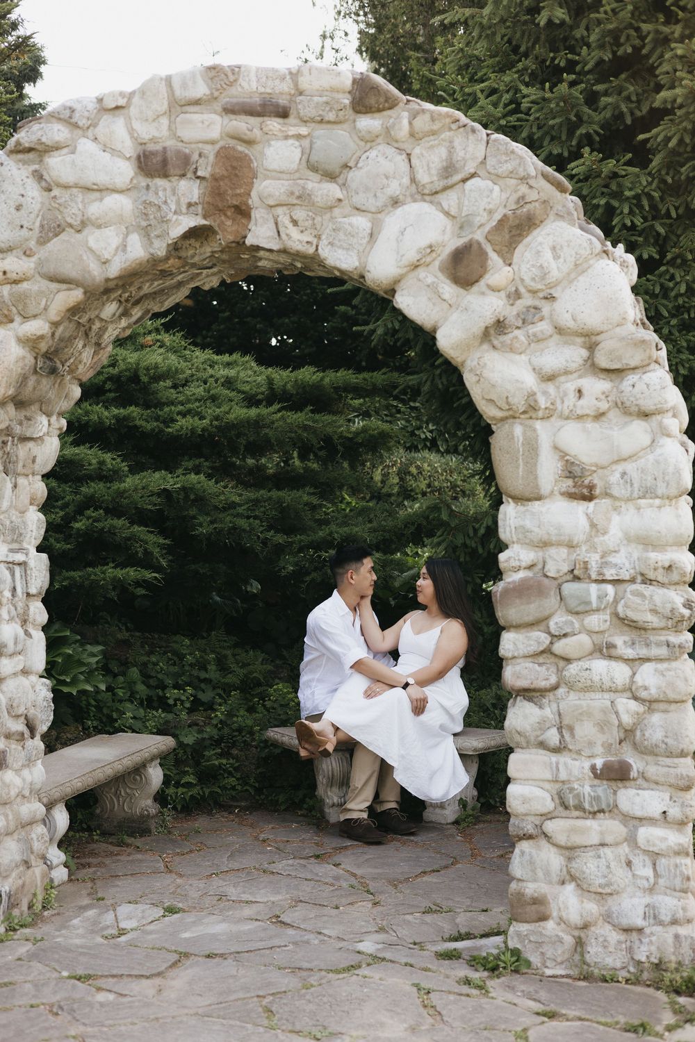 Romantic couple in white outfits sitting together under rustic stone archway in garden setting at Rockway Gardens in Kitchener Ontario
