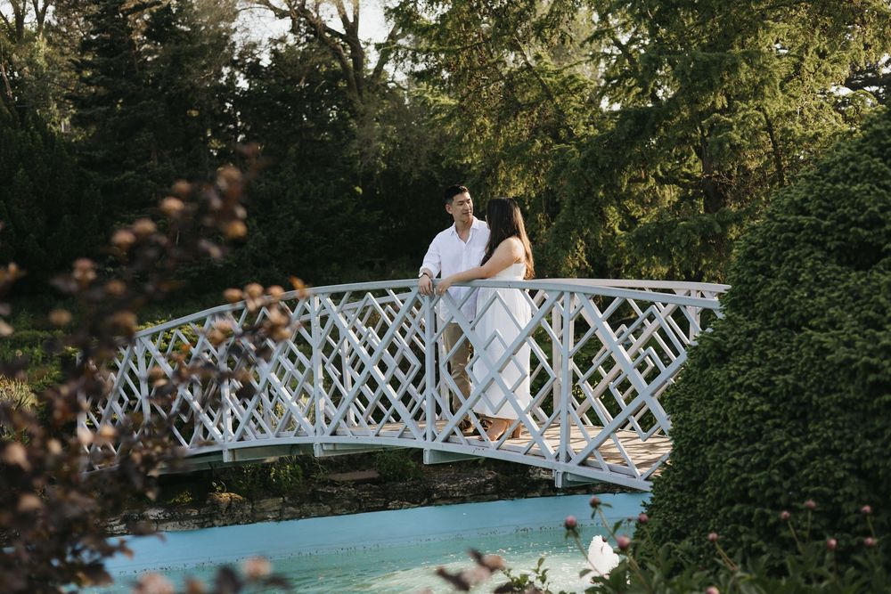 Romantic couple poses on white lattice bridge surrounded by lush greenery during wedding photography session at Rockway Gardens