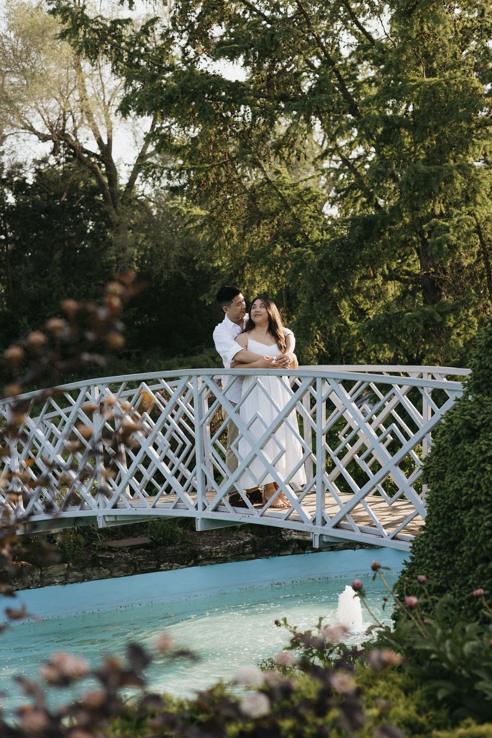 A couple stands together on a white latticed garden bridge over water surrounded by lush greenery in an outdoor setting at Rockway Gardens