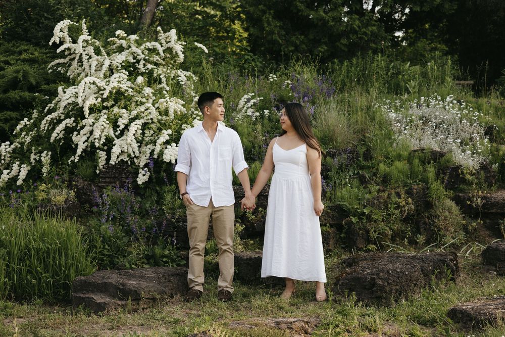 A couple in summer wedding attire walks hand in hand through a lush garden with blooming white flowers in the background at Rockway Gardens
