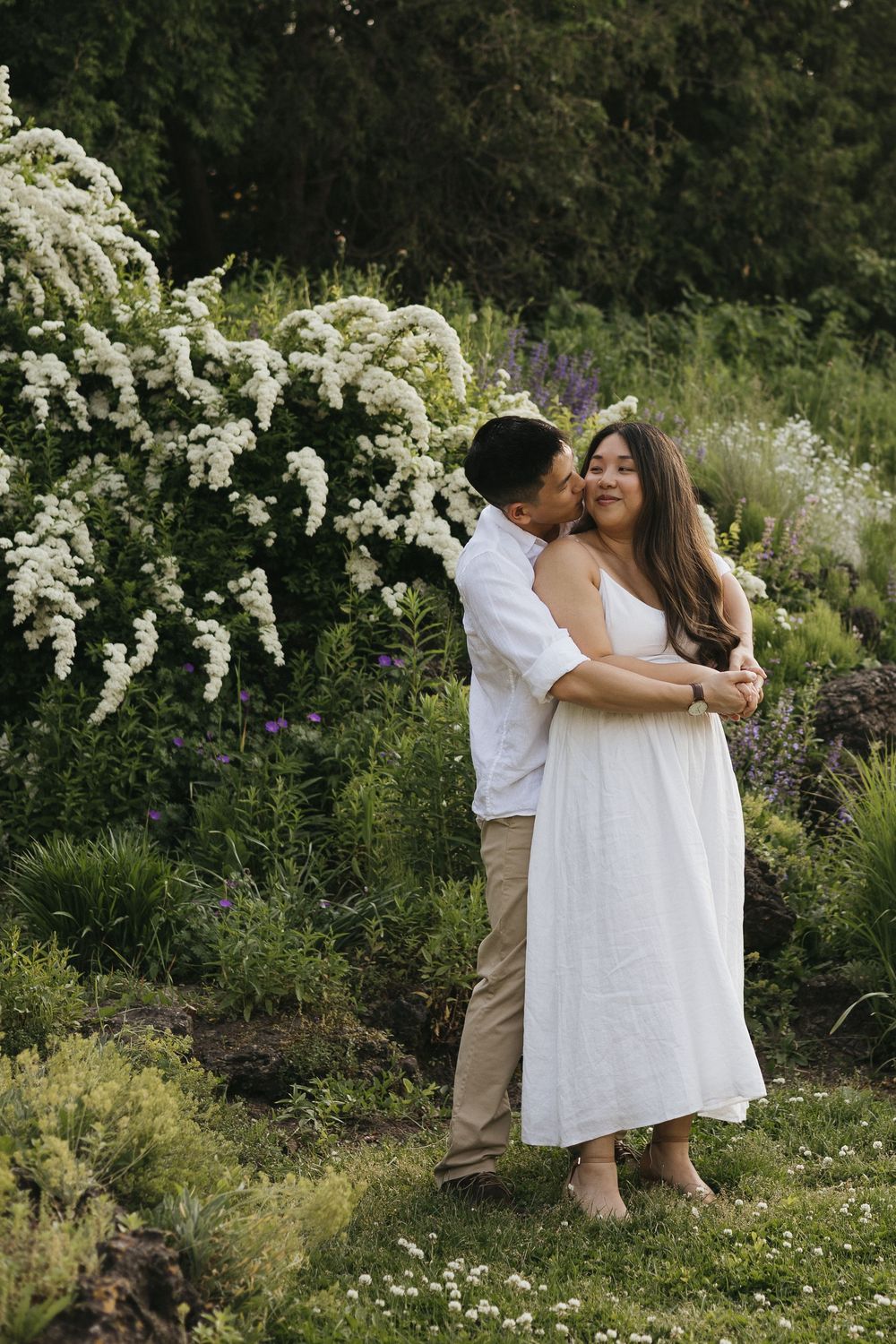 Couple embraces in romantic garden setting surrounded by blooming white flowers and lush greenery at Rockway Gardens in Kitchener