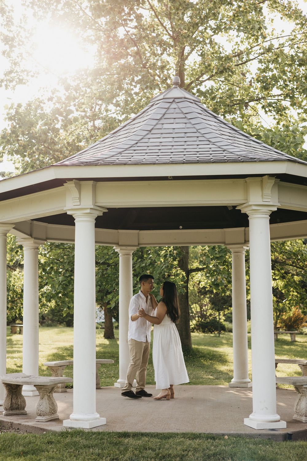 Two people share an intimate moment in a white gazebo with columns as sunlight filters through trees in the background at Rockway Gardens