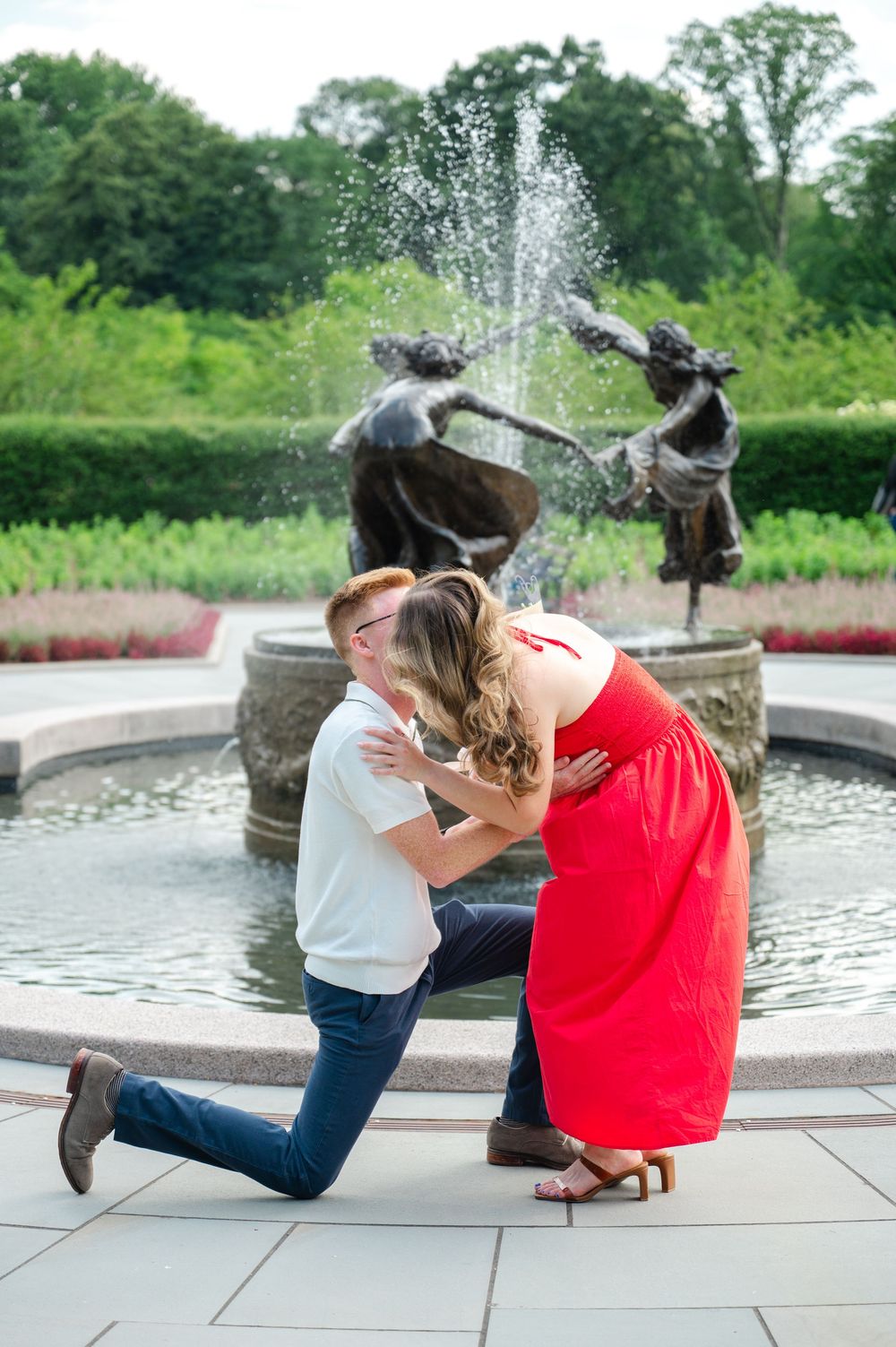 A romantic proposal moment in the Conservatory Gardens of Central Park with a woman in a red dress and a man on one knee.