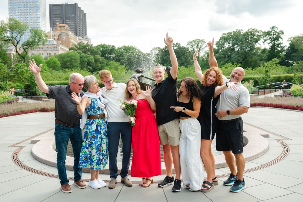 Family cheers to celebrate a couple's new engagement in Central Park, NYC. Captured by best NYC proposal photographer.
