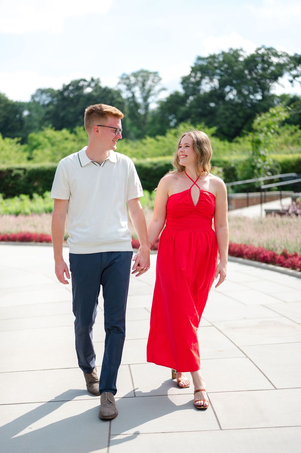 A couple enjoys a sunny walk together, with one wearing a white polo and the other in a flowing red dress.