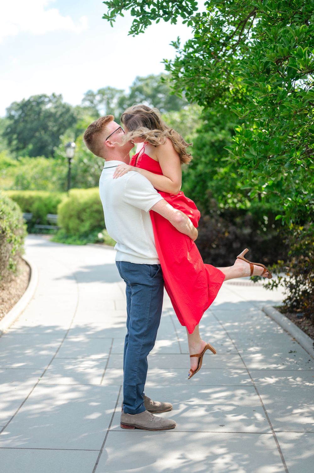 A couple embraces joyfully in Central Park, with the woman in a flowing red dress being lifted in celebration of their new proposal.