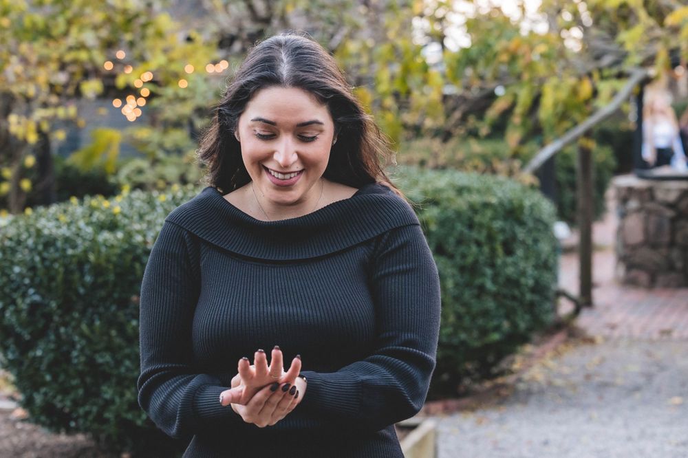 A woman in black laughs with joy while looking at her hand in an outdoor garden setting.