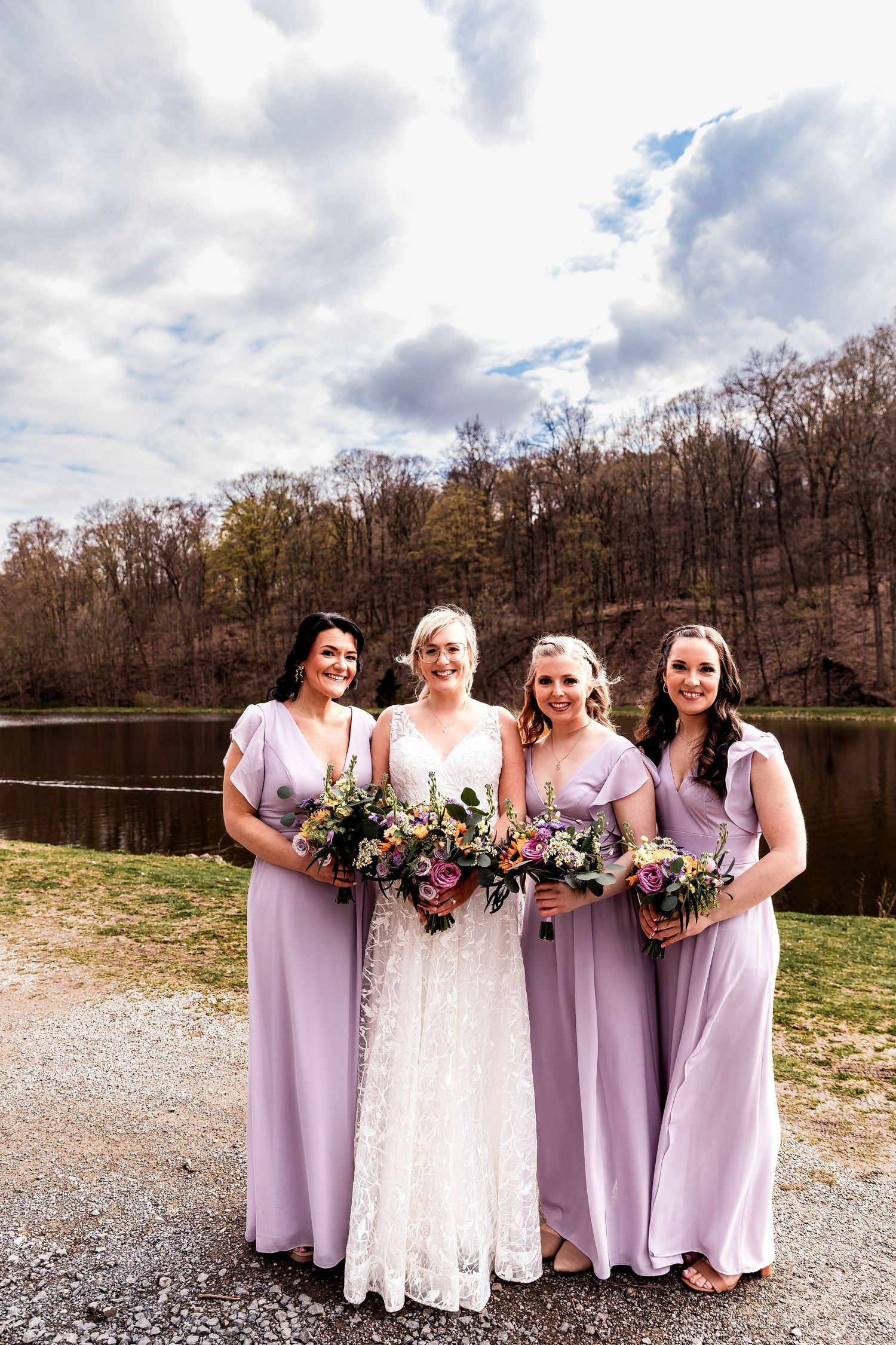 A bridal party in lilac purple bridesmaid dresses and white wedding gown pose by a serene lake on a cloudy day.