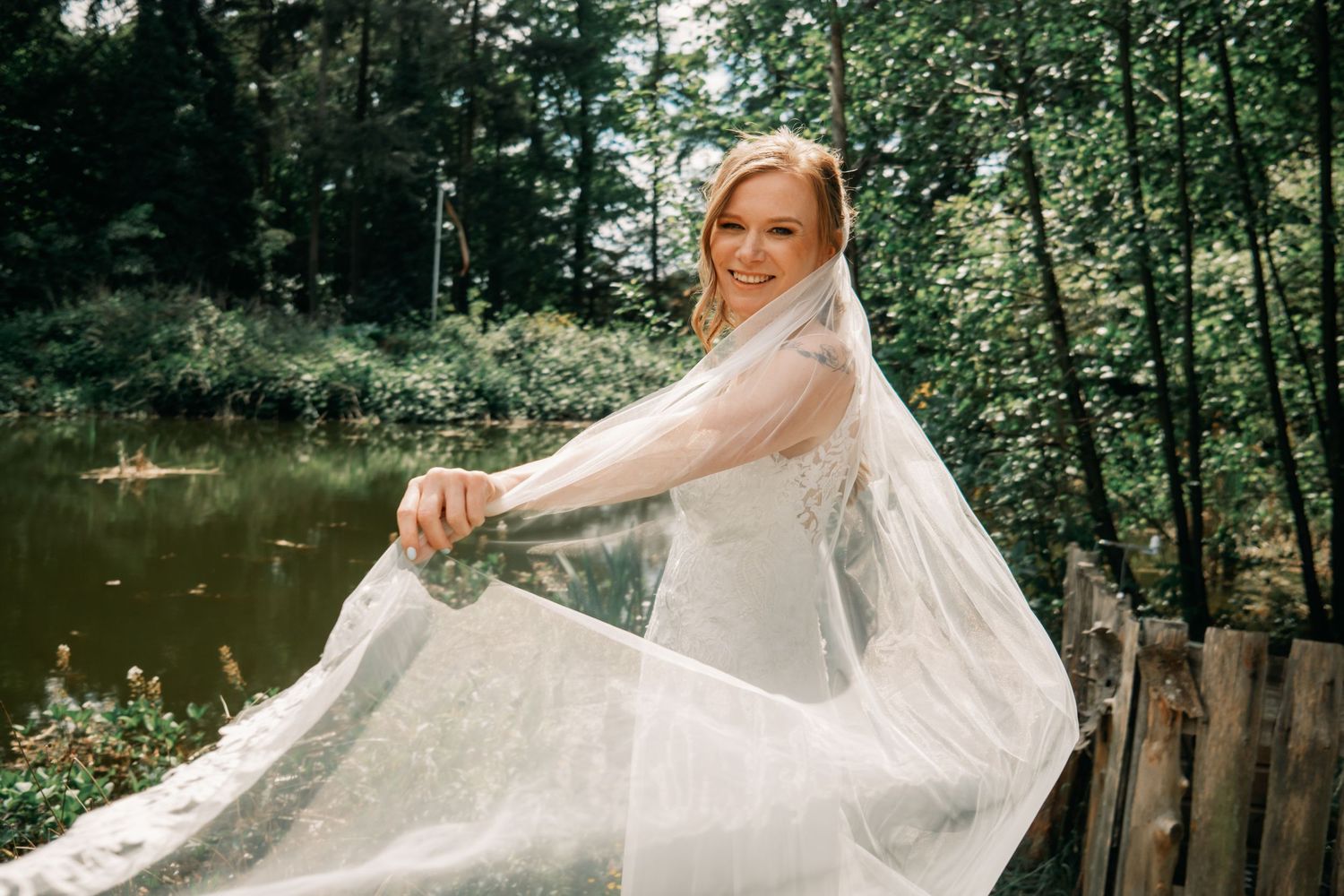 A bride in a white wedding dress and veil poses gracefully by a pond surrounded by lush greenery.