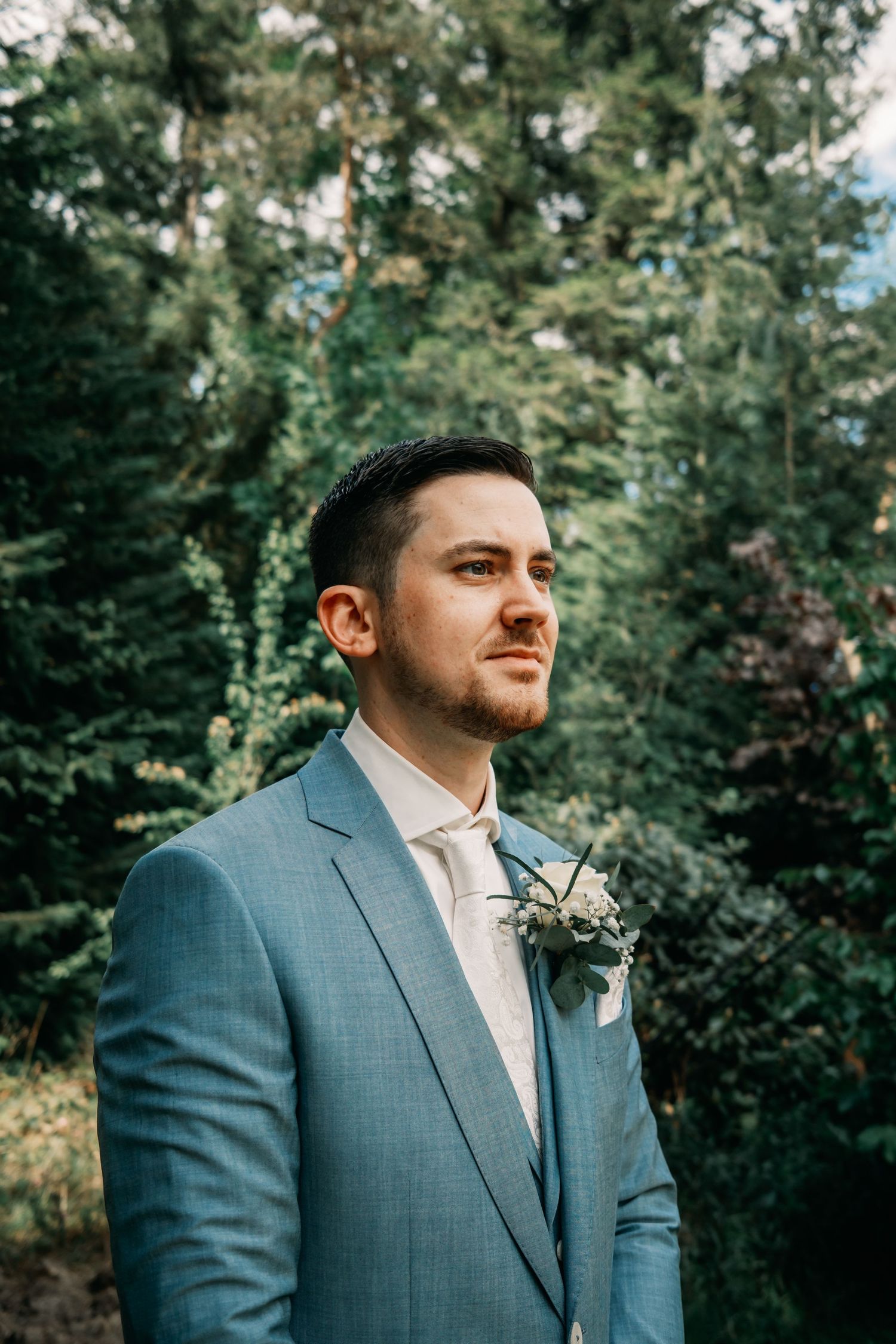 A groom in a light blue suit and white tie stands thoughtfully against a wooded outdoor backdrop during a wedding.