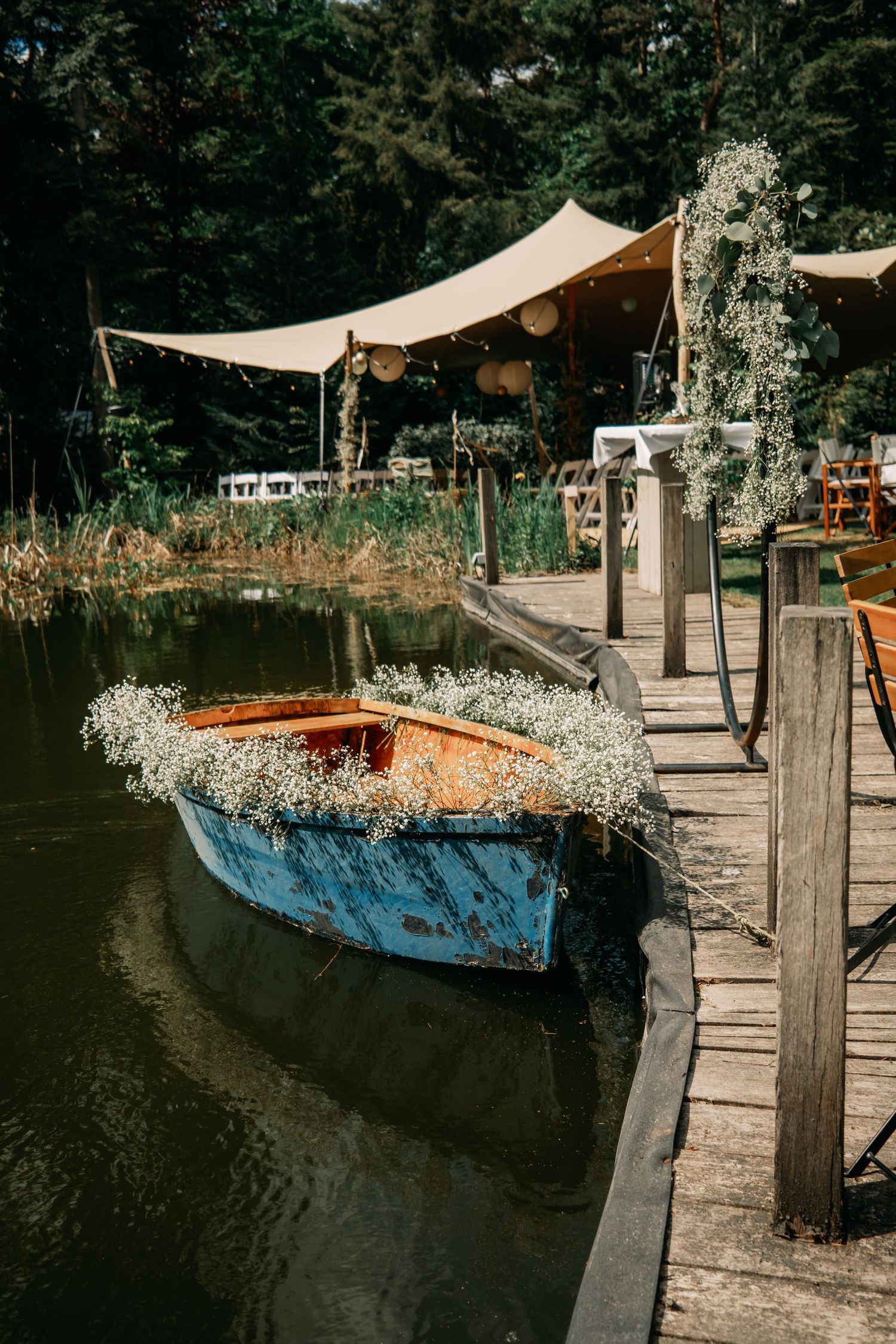 A blue wooden boat decorated with baby's breath flowers is docked by a wooden pier with a canopy tent in the background.