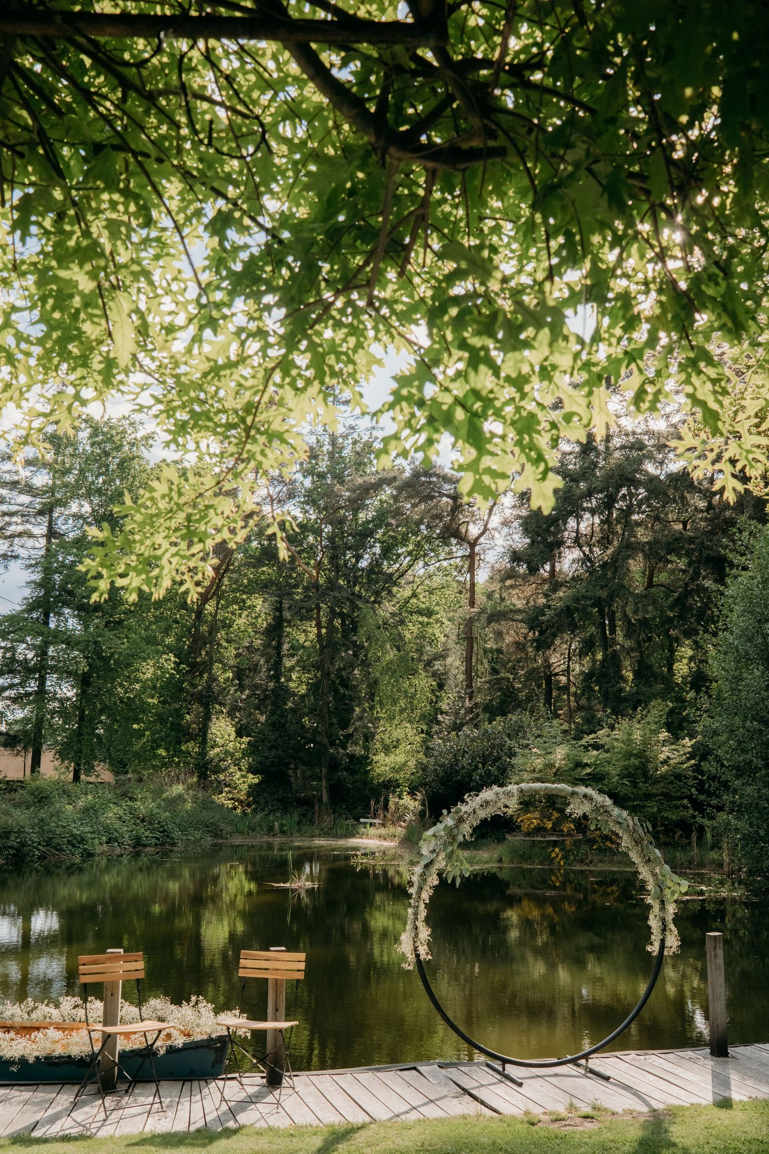 A decorative circular wedding arch stands on a wooden dock overlooking a serene pond surrounded by lush green trees.
