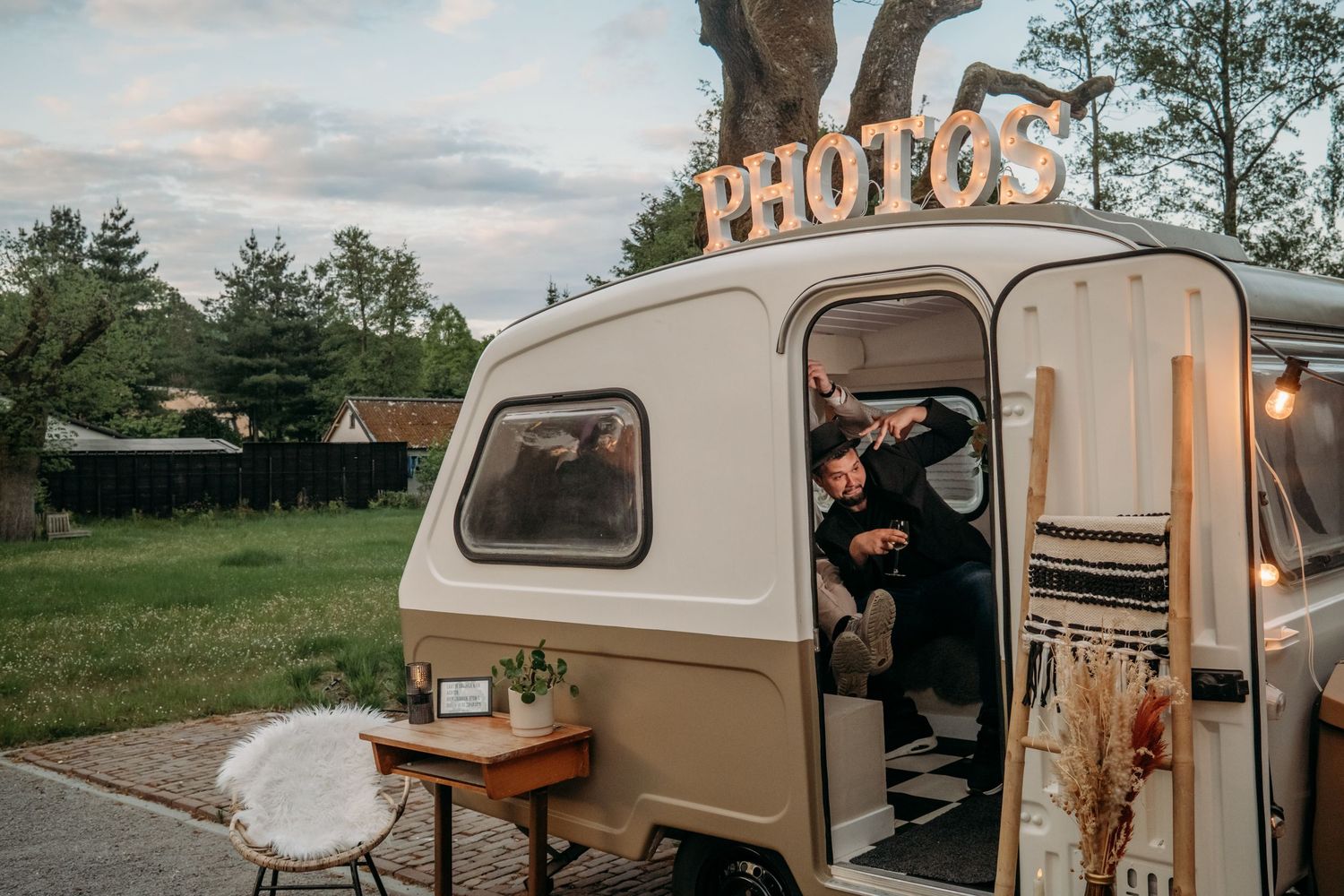 A vintage photo booth trailer with illuminated PHOTOS sign and warm lighting at outdoor event during sunset.