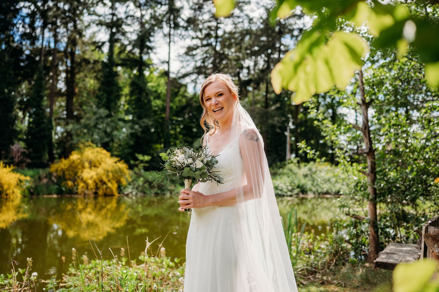 A bride in a flowing white dress and veil stands by a sunlit pond surrounded by lush garden greenery and trees.