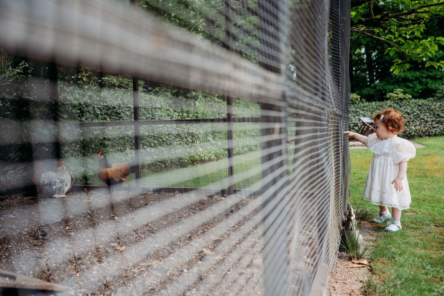 A small figure in a white dress stands near a reflective memorial wall with trees visible in the background.