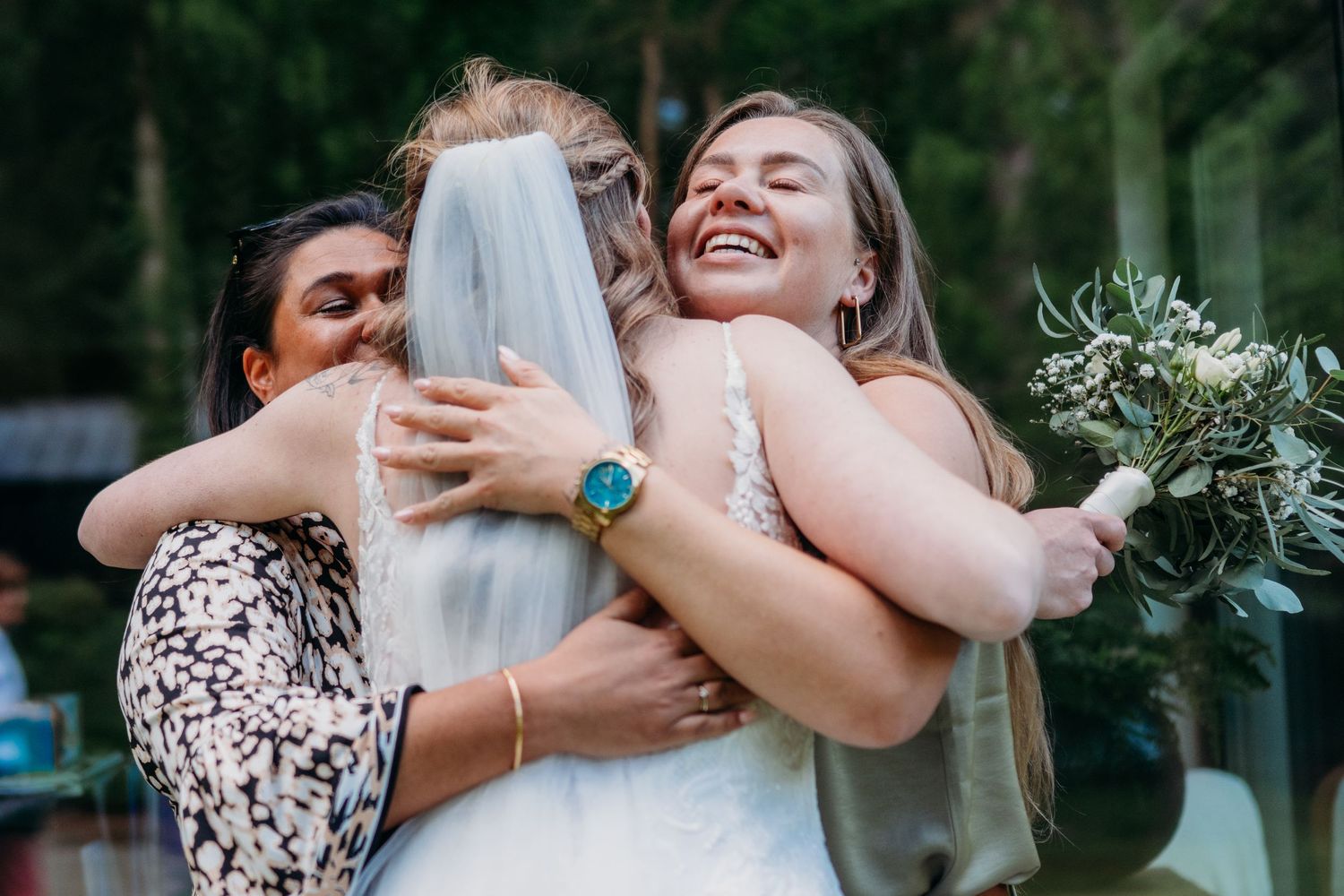 Three people share an emotional group hug at an outdoor wedding celebration.