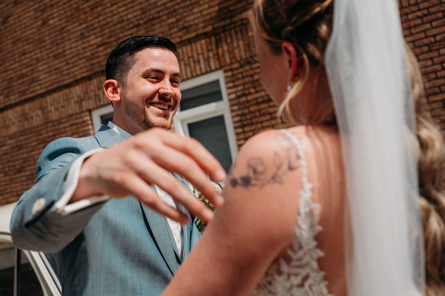 A joyful wedding moment between the couple standing against a brick wall in bright sunlight.