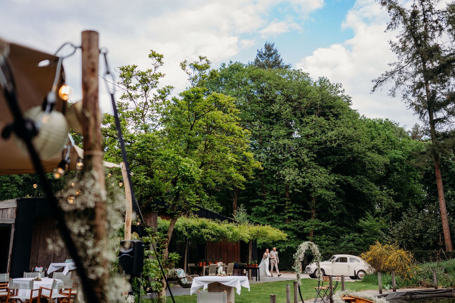 White outdoor wedding reception setup in a garden with trees and greenery in the background.