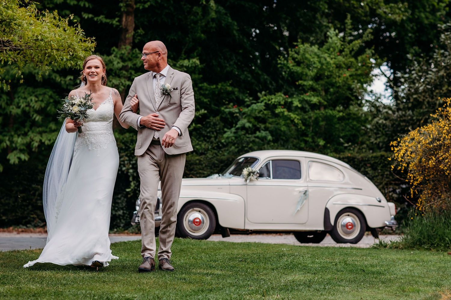A couple in wedding attire walking on a lawn with a vintage cream-colored Volvo car in the background.