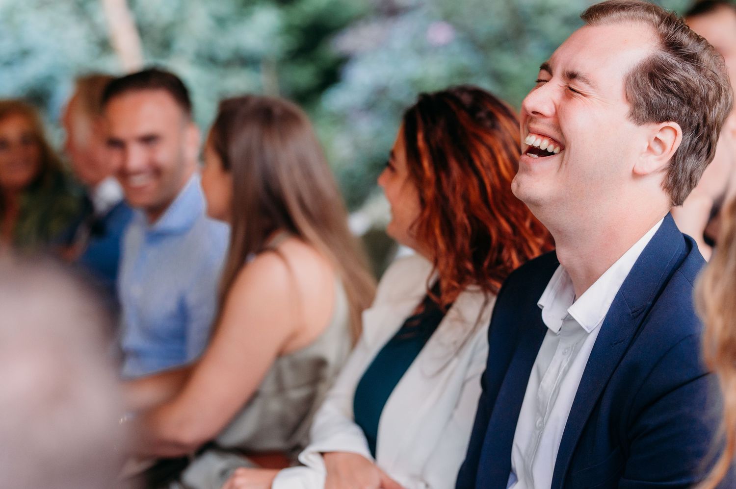 A person in a navy suit laughs joyfully during an outdoor social gathering as others converse in the background.