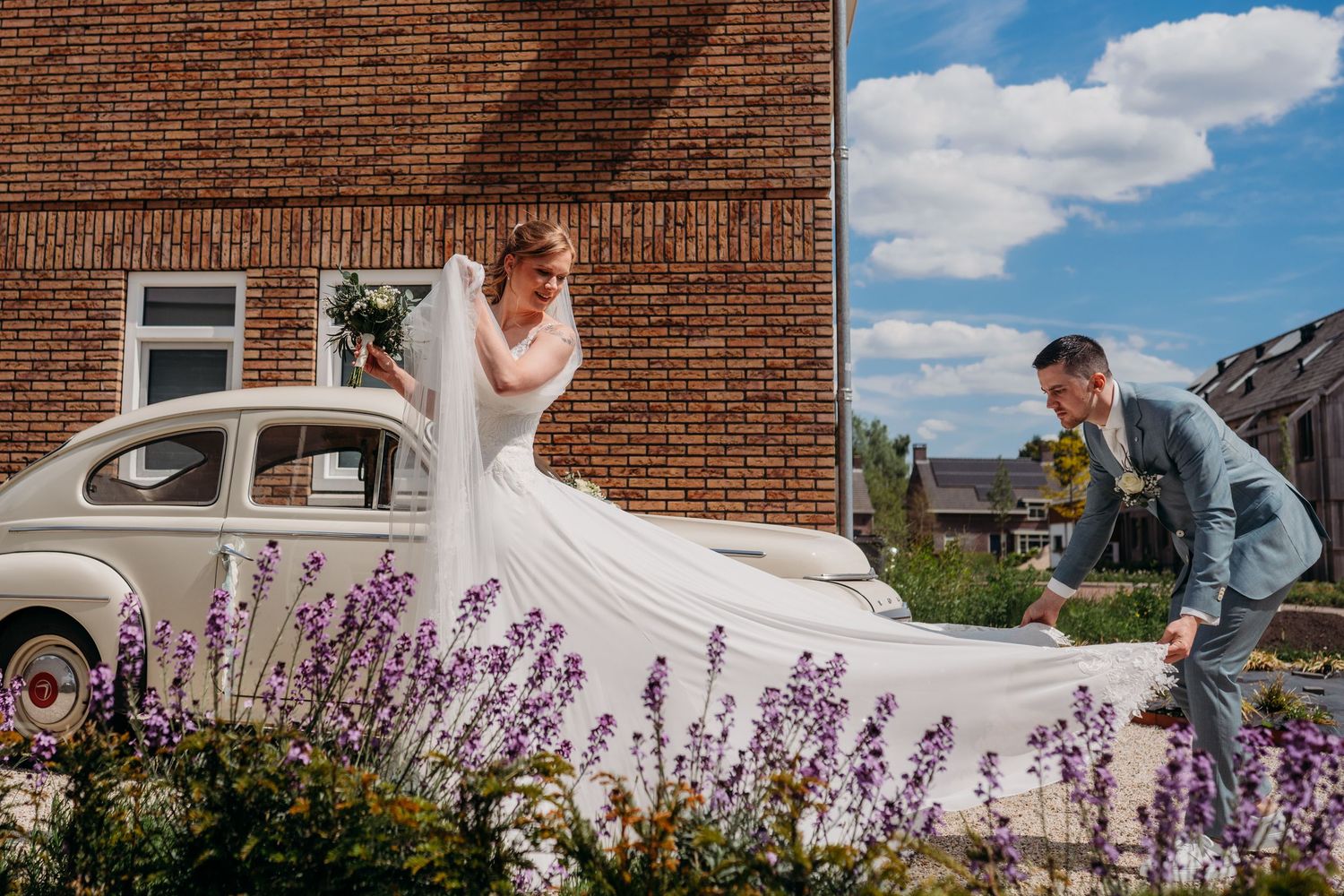 A bride in white dress stands near vintage car with lavender flowers while someone leans in playfully.
