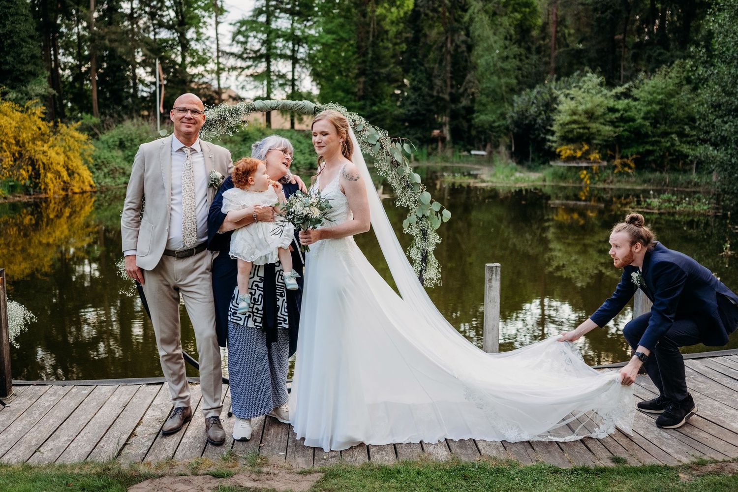 A joyful wedding party stands on a wooden dock by a tranquil pond surrounded by lush greenery.