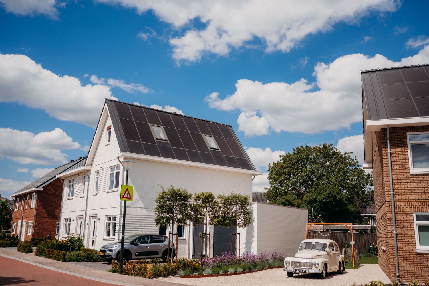 Modern white house with solar panels on sloped roof and parked cars in front under bright blue sky with puffy clouds.