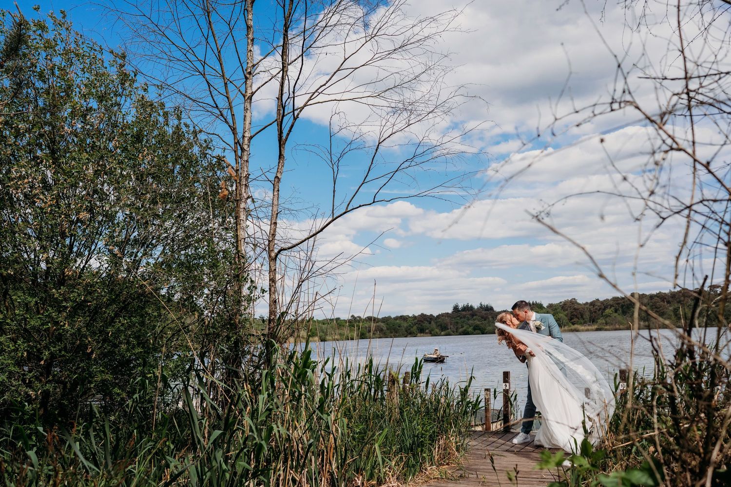 Wind blows a white dress during an outdoor photo shoot by a scenic lake with tall grass and bare trees.