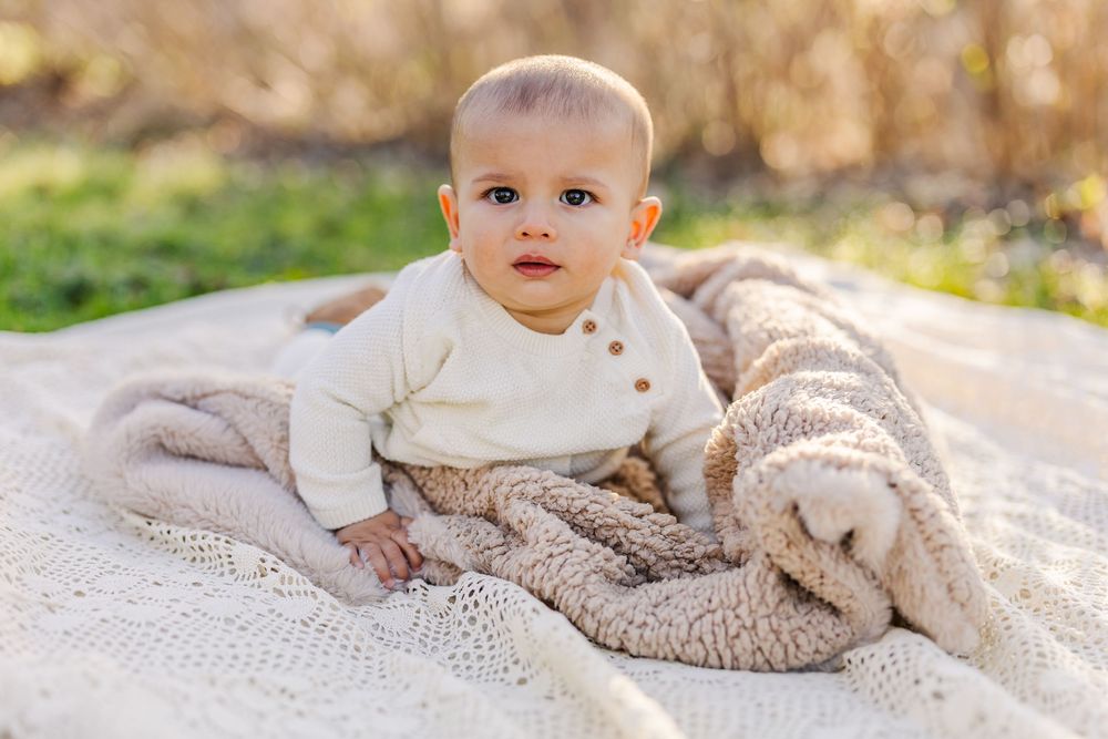 Baby in a cream colored sweater sits on a cozy knit blanket in warm outdoor lighting.