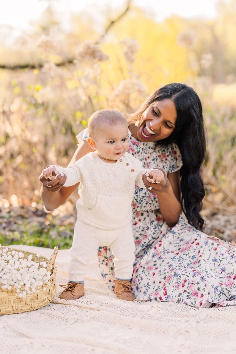Baby takes first steps on blanket while parent in floral dress provides support during outdoor photo session.