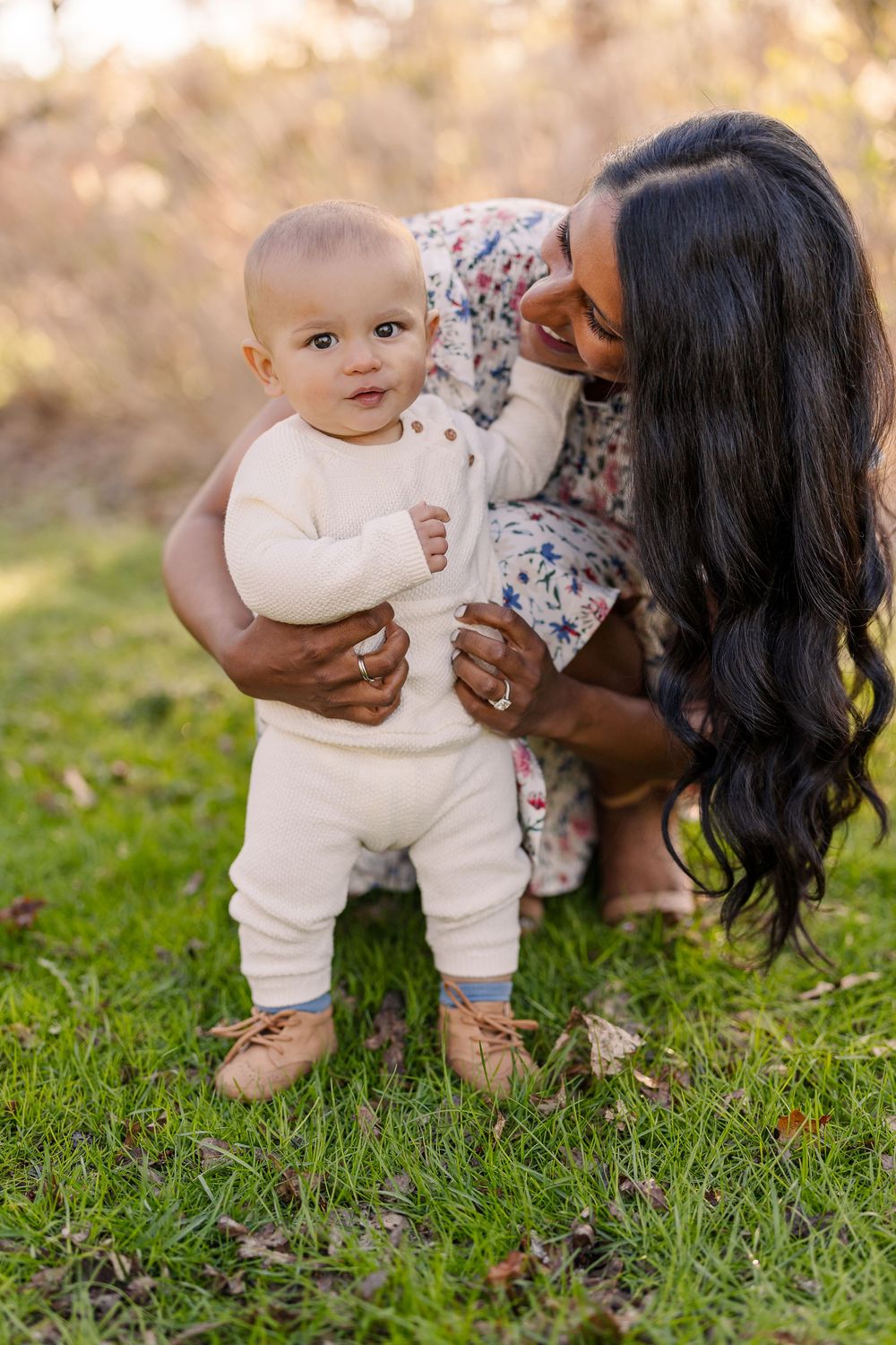 Sweet mother-child moment outdoors featuring a baby in white outfit and booties standing on grass during golden hour.