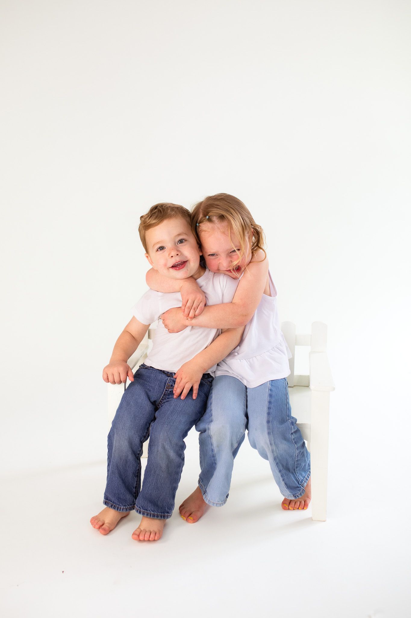 Two children in white shirts and jeans sit together barefoot on a white cube against a light background.