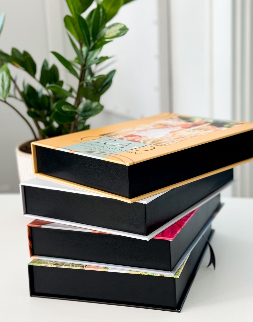 A stack of black hardcover books with colorful spines arranged on a white surface near a green potted plant.