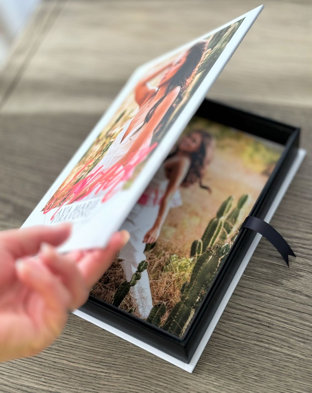 Hand holding a printed photo album page showing artistic wedding photographs on a wooden surface.