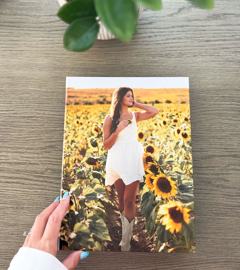 Someone in a white sundress holds up a photo while standing in a vibrant sunflower field at golden hour.