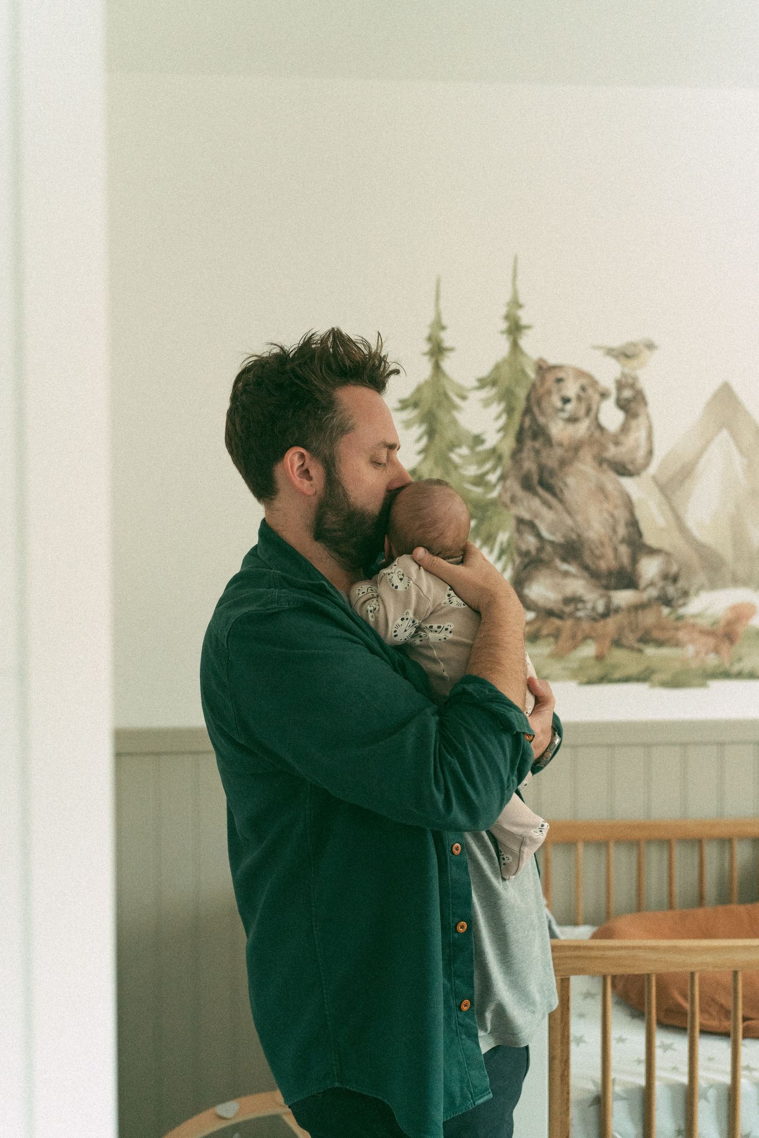 A tender nursery moment with parent holding an infant in a nursery with woodland-themed artwork.