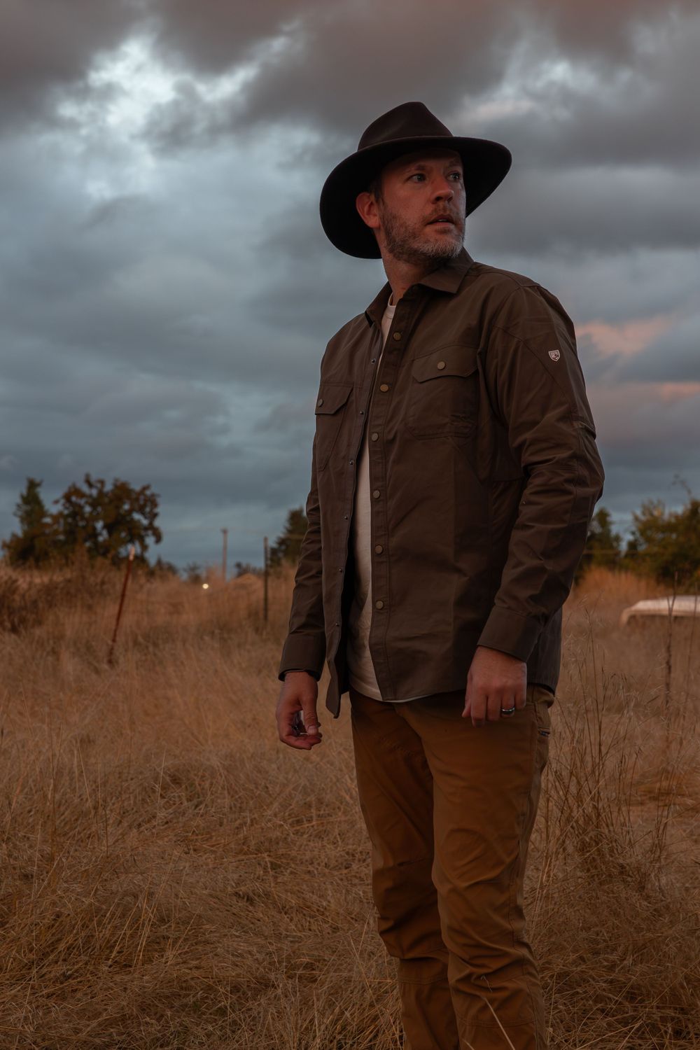 Person in brown jacket and wide-brim hat stands in golden field during sunset.